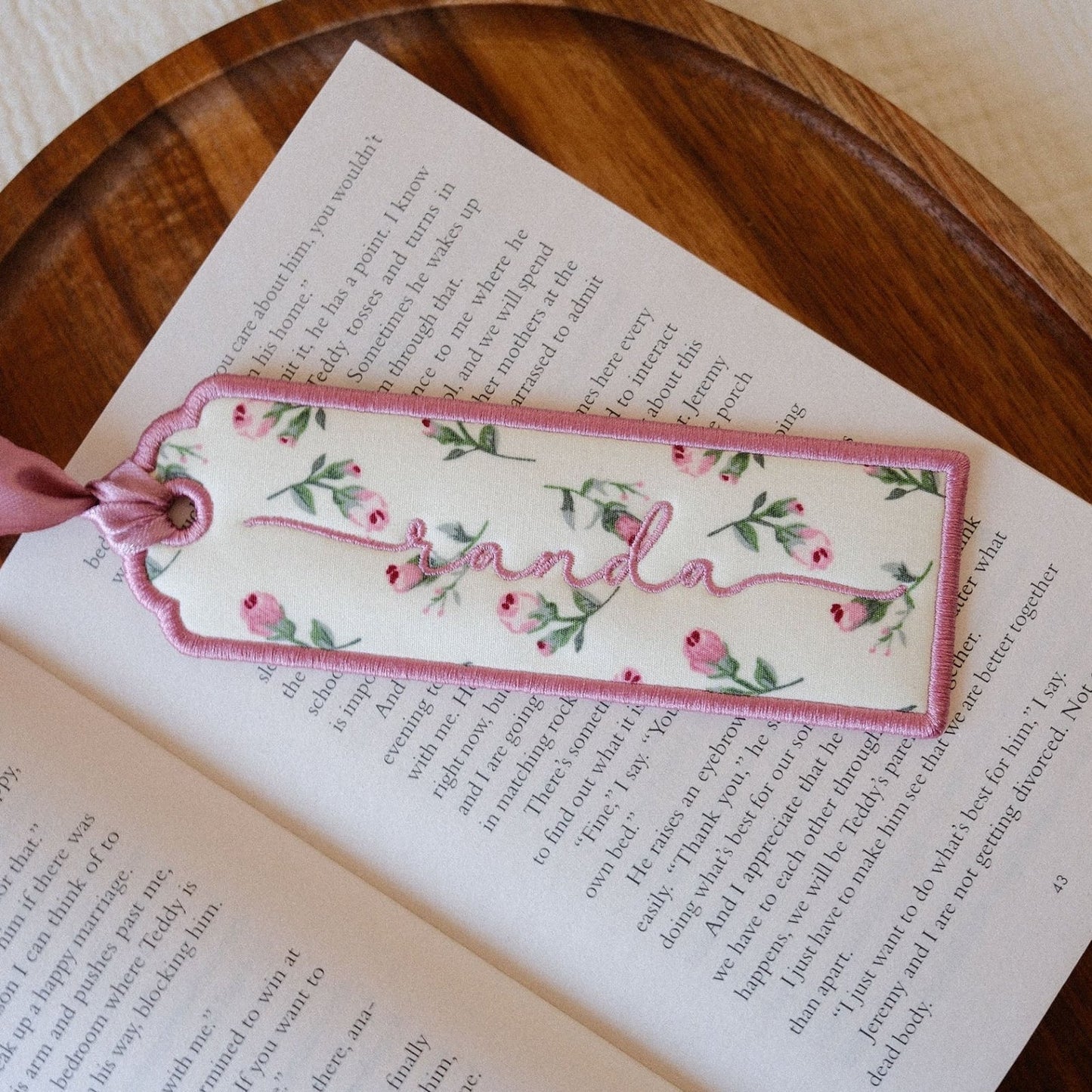 A rectangular fabric bookmark with a pink border and embroidered floral pattern, tied with a pink ribbon, resting on an open book on a wooden tray.