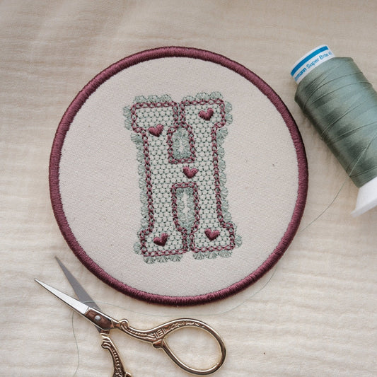 Personalised embroidered monogram coaster with lace effect letter H in plum thread and sewing accessories nearby