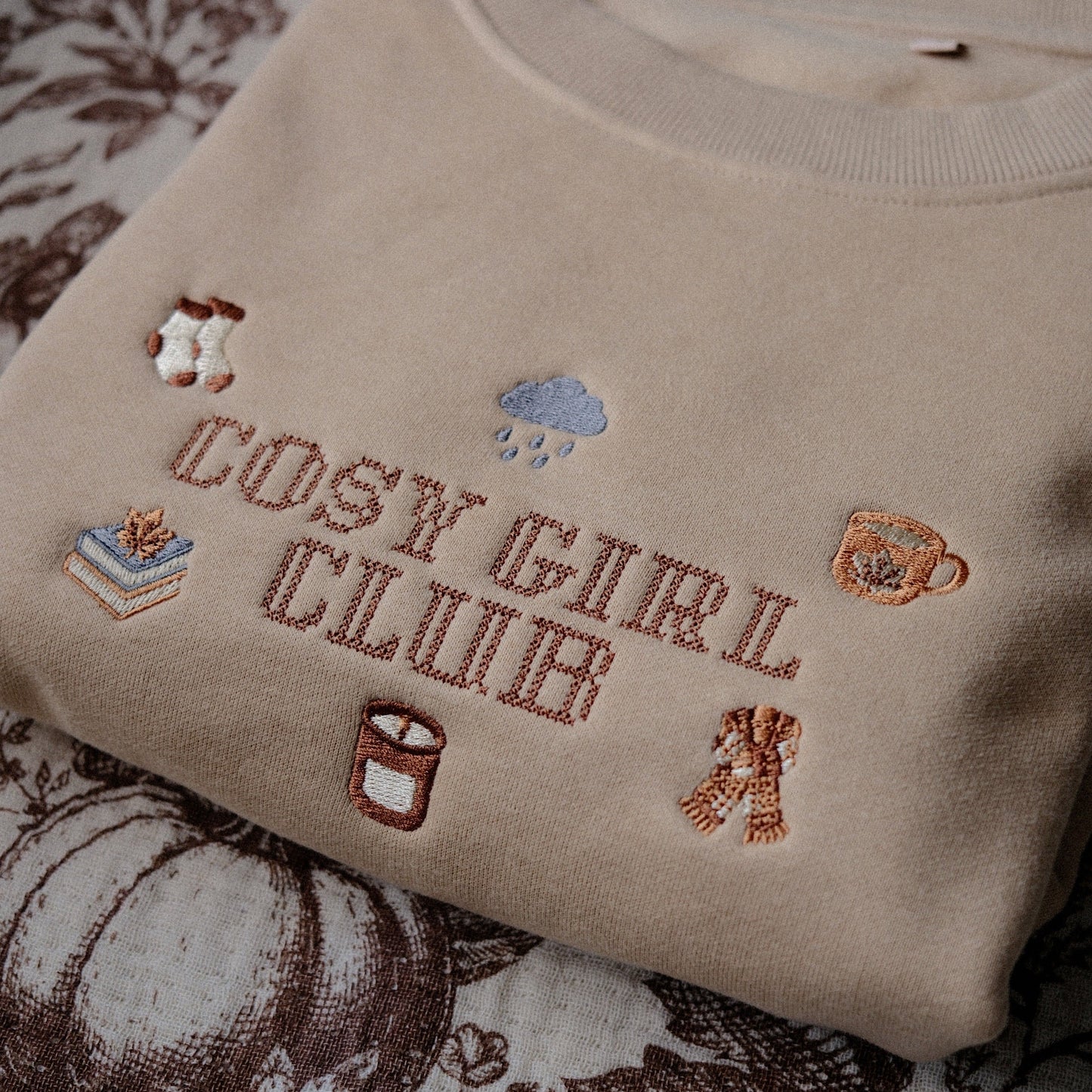 A beige crewneck sweatshirt with 'COSY GIRL CLUB' embroidered in brown across the chest, surrounded by small autumn-themed embroidery motifs, laid flat on a brown and cream floral quilted blanket.