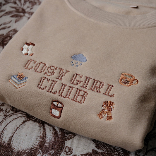 A beige crewneck sweatshirt with 'COSY GIRL CLUB' embroidered in brown across the chest, surrounded by small autumn-themed embroidery motifs, laid flat on a brown and cream floral quilted blanket.