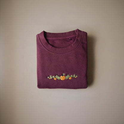 A folded crewneck sweatshirt in a deep burgundy colour, featuring an embroidered autumn vine design with orange pumpkins and green leaves across the lower chest, set against a plain beige background.