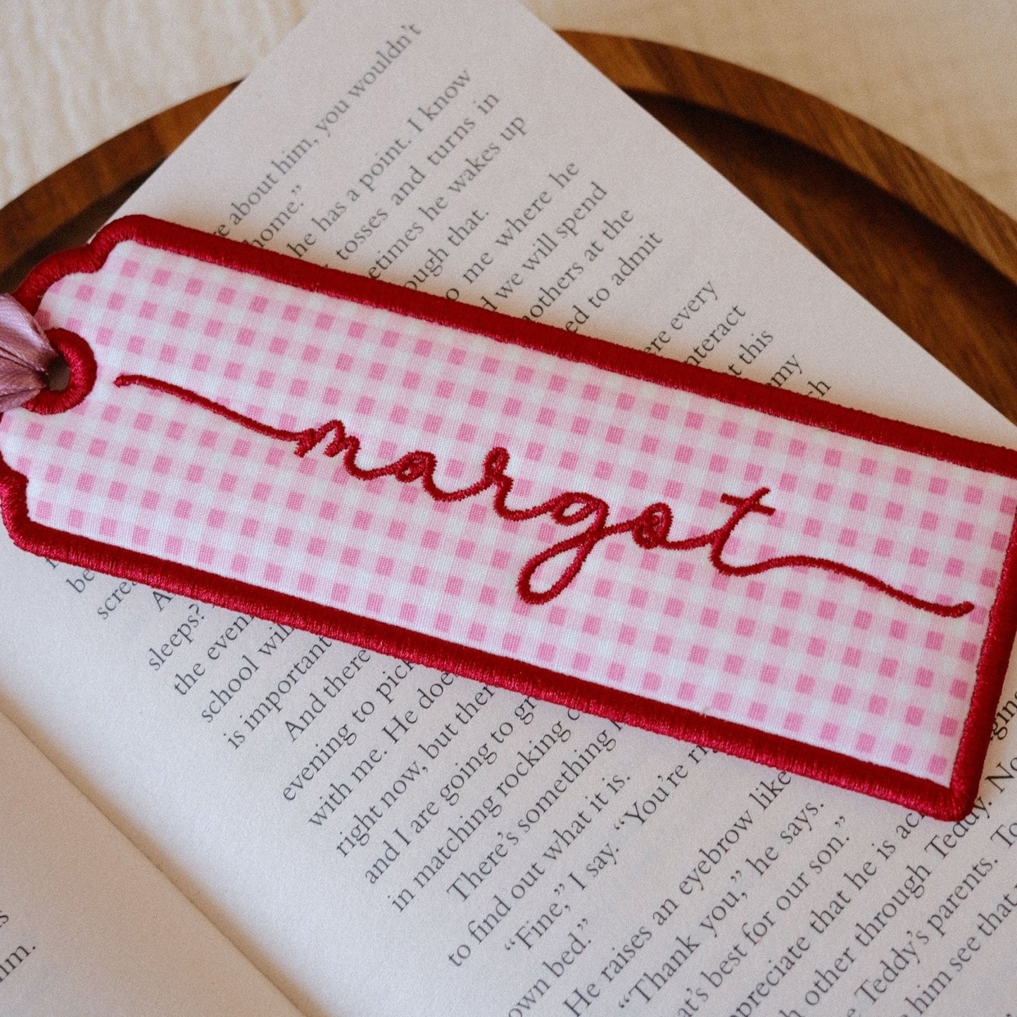 A pink and white gingham fabric bookmark with red embroidered script reading 'margot' and a red stitched border, placed on an open book.