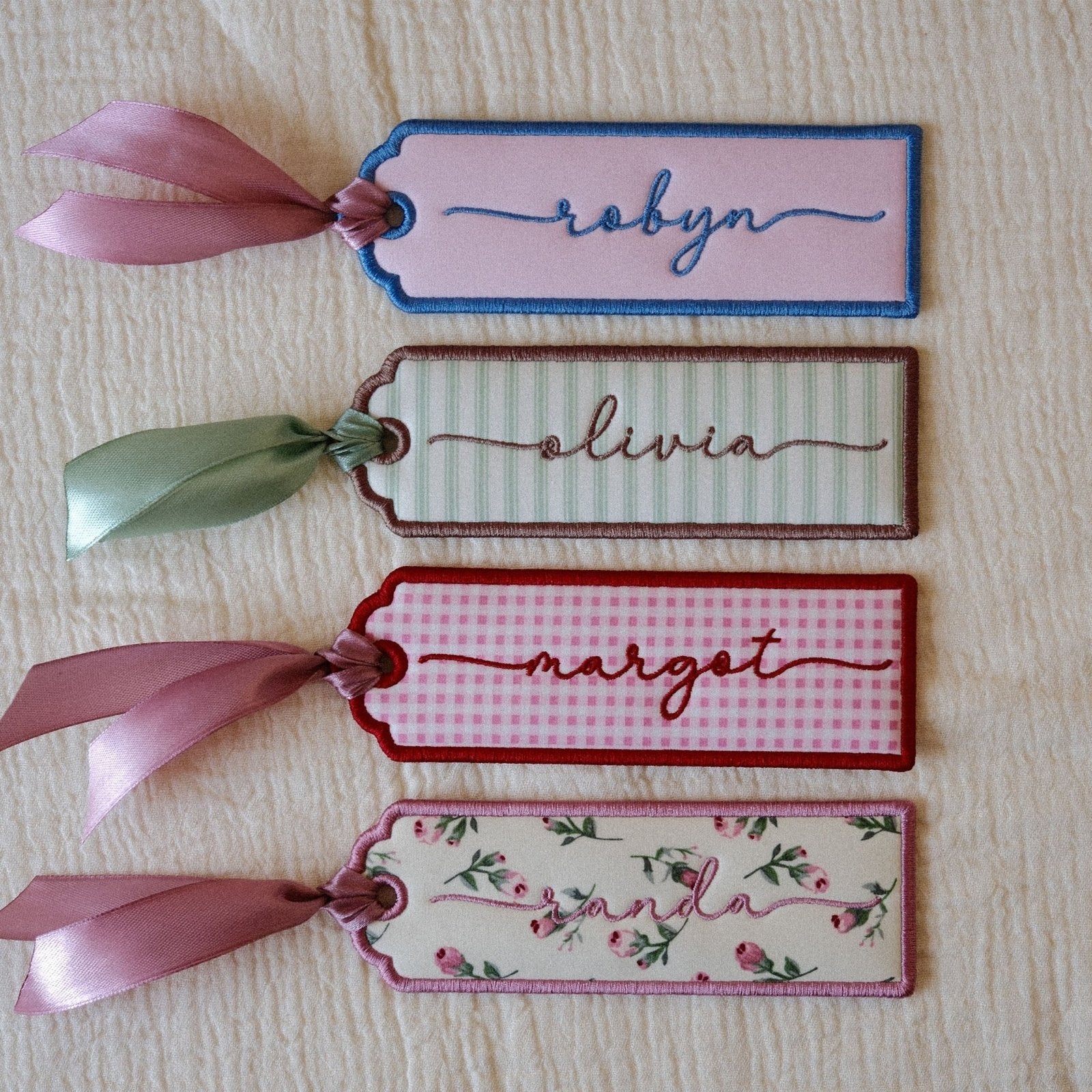 Four decorative fabric bookmarks with ribbon ties are arranged vertically on a textured cream fabric background. The tags feature different patterns and colors: light pink with blue border and the name 'robyn', cream with green border and the name 'olivia', pink with red border and the name 'margot', and floral with pink border and the name 'randa'.
