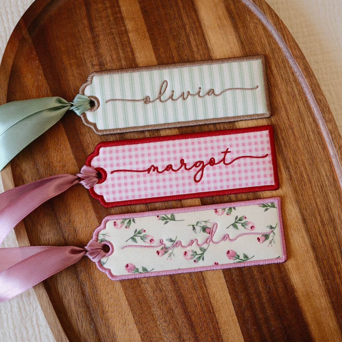 Three decorative personalised bookmarks with ribbons, arranged on a wooden serving board over a light fabric background. The tags feature striped, gingham, and floral patterns in soft pastel colours.