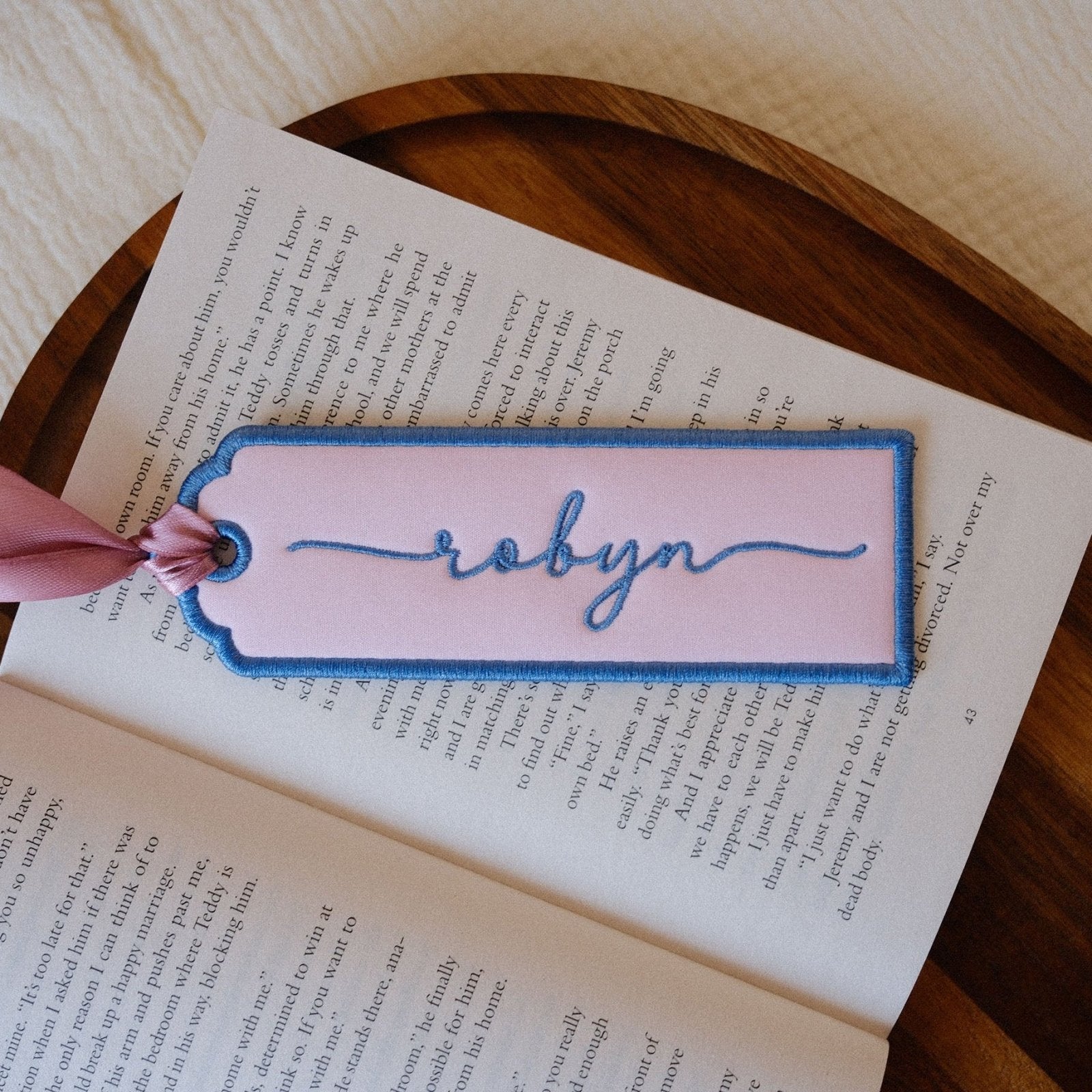 A pink embroidered bookmark with blue border and the name 'robyn' stitched in blue script, resting on an open book with a wooden tray in the background.