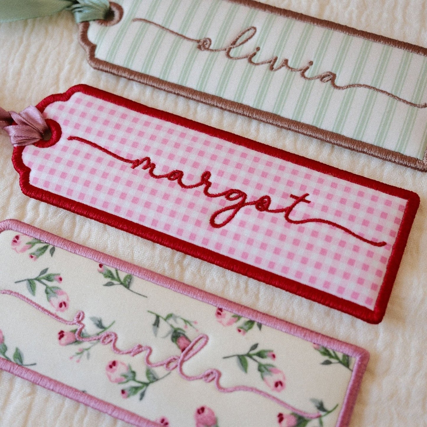 Three personalized embroidered fabric tags with ribbon loops, laid flat on a textured cream fabric background. The top tag has pale green and white stripes with brown embroidery; the middle tag has a pink gingham pattern with red embroidery and border; the bottom tag has a pink floral print with green leaves and pink embroidery.
