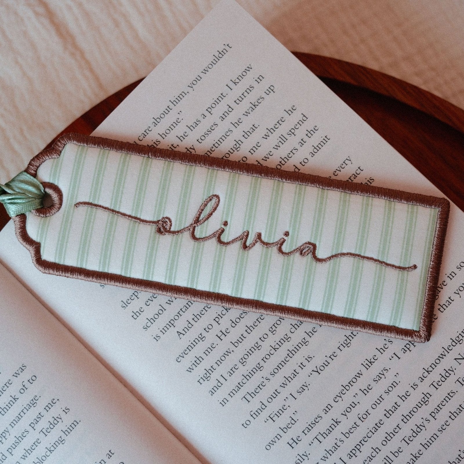 A personalized fabric bookmark shaped like a book tag, featuring green and white stripes, brown embroidered border and the name 'Olivia' in brown script, resting on an open book.