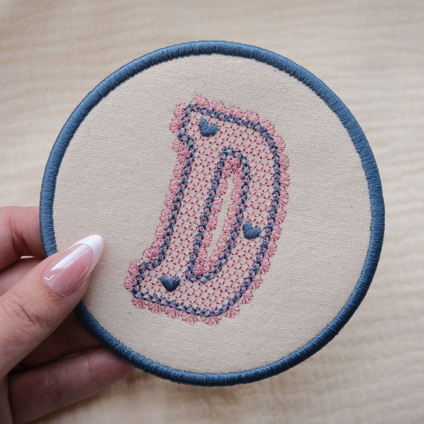 Embroidered lace initial coaster in blue and pink thread colours, held in hand