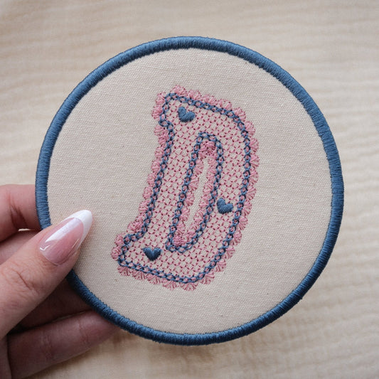 Embroidered lace initial coaster in blue and pink thread colours, held in hand