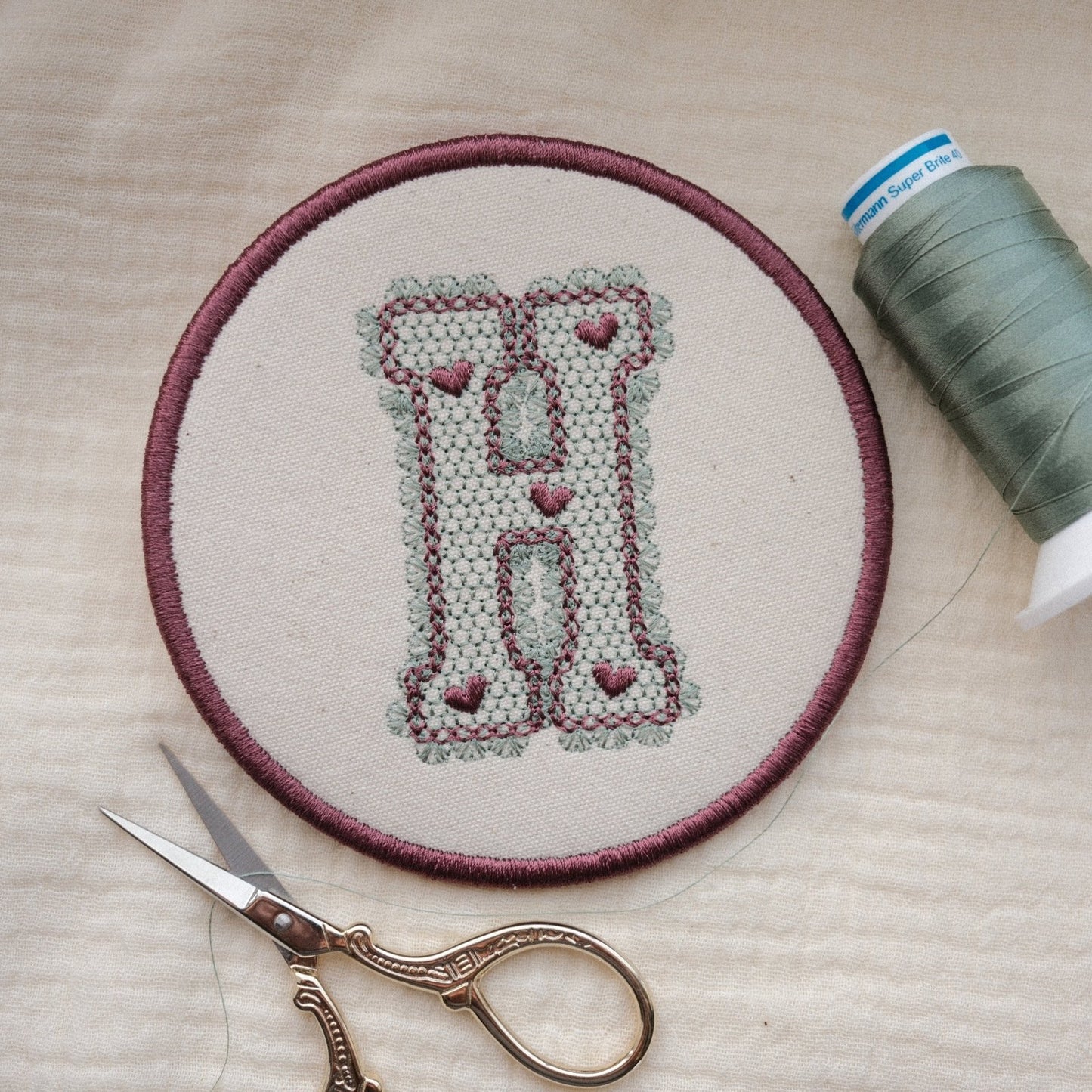 Personalised embroidered monogram coaster with lace effect letter H in plum thread and sewing accessories nearby