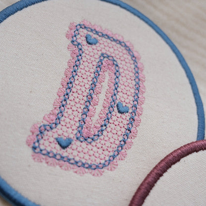 Detailed close up of lace style embroidered letter D coaster with pink stitching and blue embroidered edge